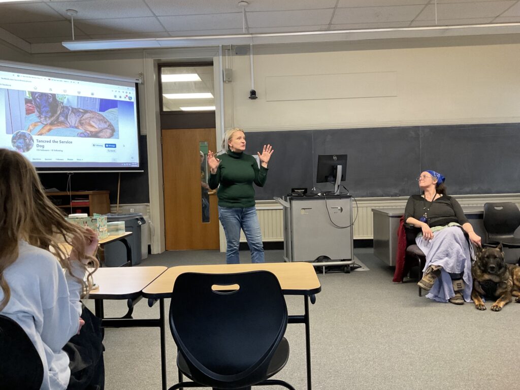 In a classroom, white female teacher, Christine Carpenter, in jeans and a green turtleneck, leads the class. A projector screen behind her has an image of a dog; next to her, a white woman in a light blue skirt is seated next to a leashed german shepard.  