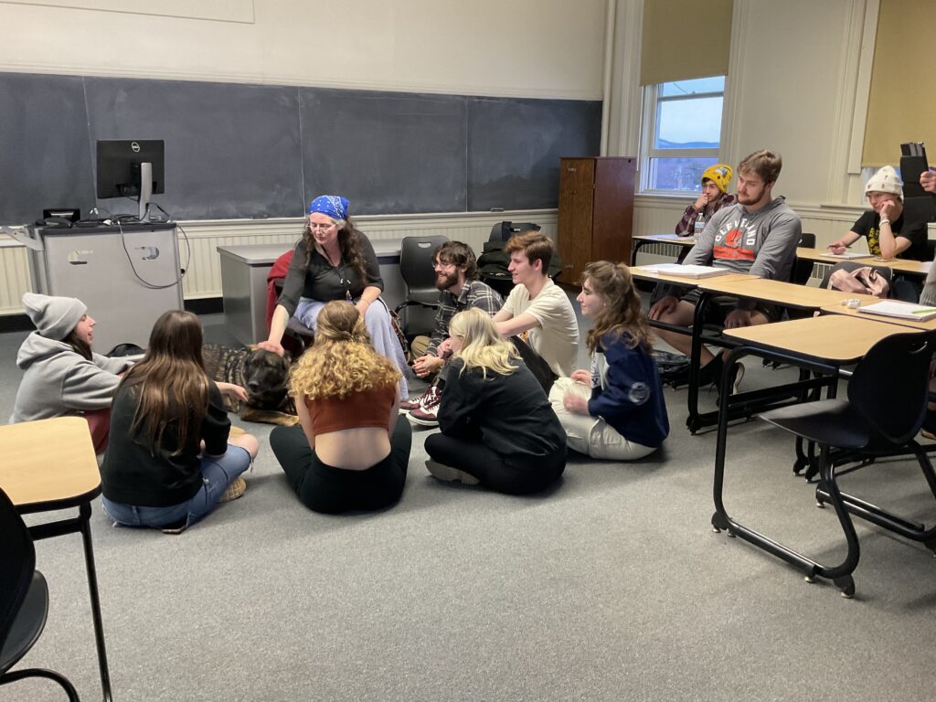 In a classroom, a white woman with long brown hair and a blue bandanna as a headscarf sits on a chair next to a German shepard, her hand on his back. Gathered around her on the floor are a number of students; a few occupy desks in the background.