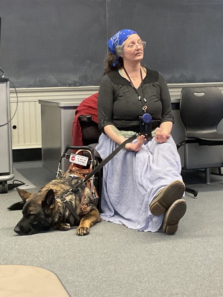 A white woman with glasses and a blue bandanna over long brown hair sits in a classroom, holding the leash of a German Shepard service dog.